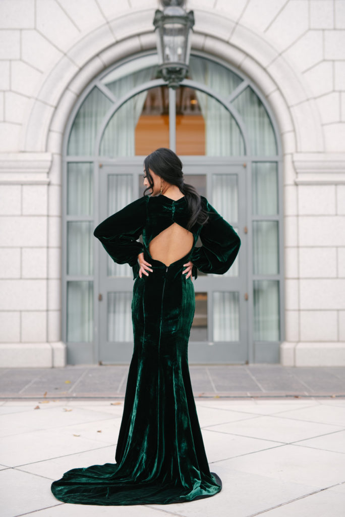 A woman in a beautiful long sleeve green velvet dress. She is turned away from the camera to show off the open back detail this dress has. Her head slightly turned to the floor.