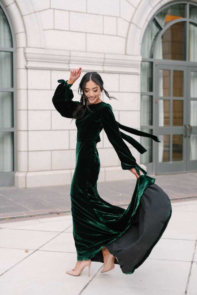 A woman in a beautiful long sleeve green velvet dress. She holds the end of her dress in one hand and the other in the air as she twirls around for the camera. Her smile is bright and she is rockin the nude tall heels!