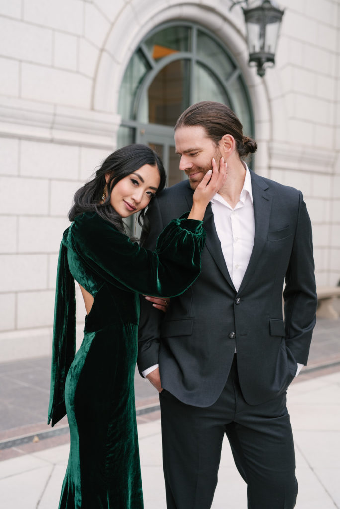 A woman in a beautiful long sleeve green velvet dress holding the man's face. The man is in a dark grey suit. He smiles at her as she smiles at the camera. This picture is serene and calm.