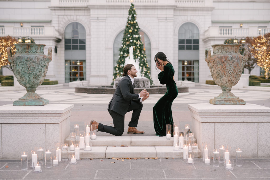 Man down on one knee proposing to a woman in a beautiful long green velvet dress. They are surrounded by white candles lit in vases. The Christmas tree is blurred but still seen in the background. This is a marriage proposal in action.