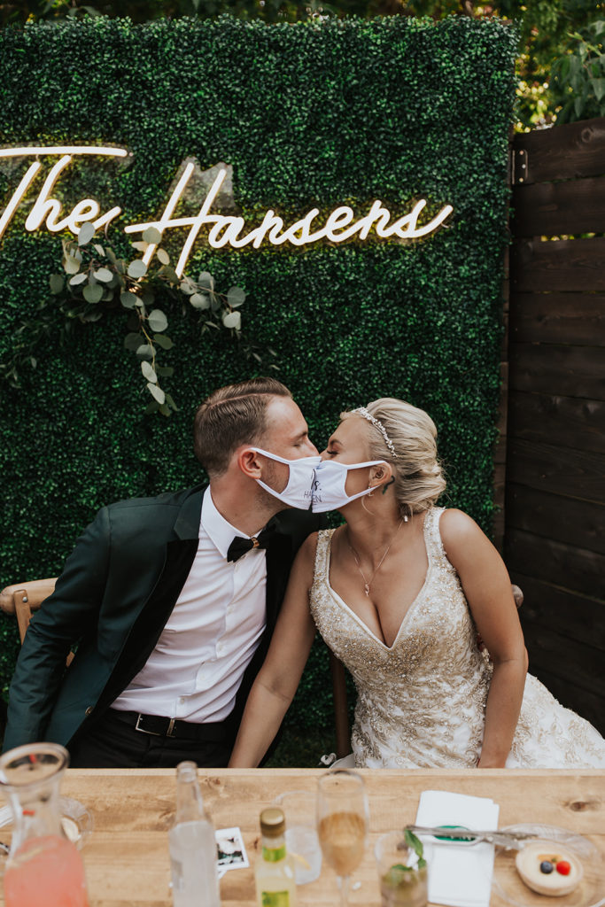 This photo is of a bride and groom in their COVID-19 mask and they are kissing. They are seated at their reception table, and behind them is a greenery wall with a glowed up sign saying "The Hansen's" 