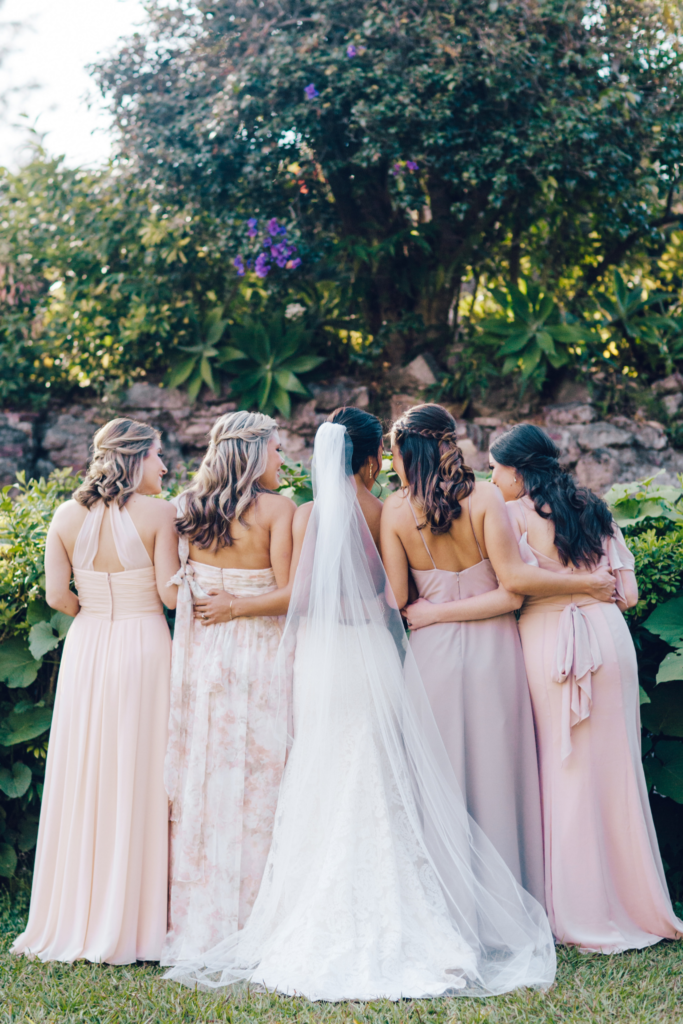 This non-traditional wedding has 4 bridesmaids all in different style dresses. The color palette is pink but they wear different styles and shades of the color palette. Their backs are facing away from the camera, arms wrapped around each other with the bride in the middle. The bride is in a white dress and wearing a long white veil. 