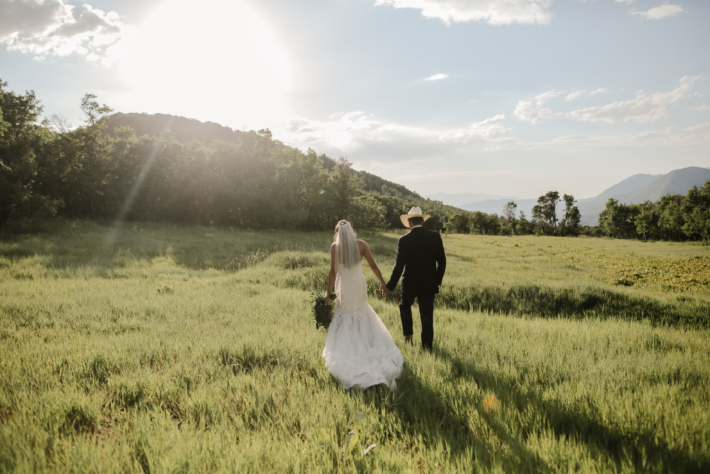 A woman in a white wedding dress with a train at the back of the dress. She also wears a white veil and holds a bouquet on one hand. Her other hand is holding the husband's hand. The male husband wears a black suite and a cowboy hat. The scenery is the couple walking through green grass and a mountain hill. The sun is shining bright with blue skies.