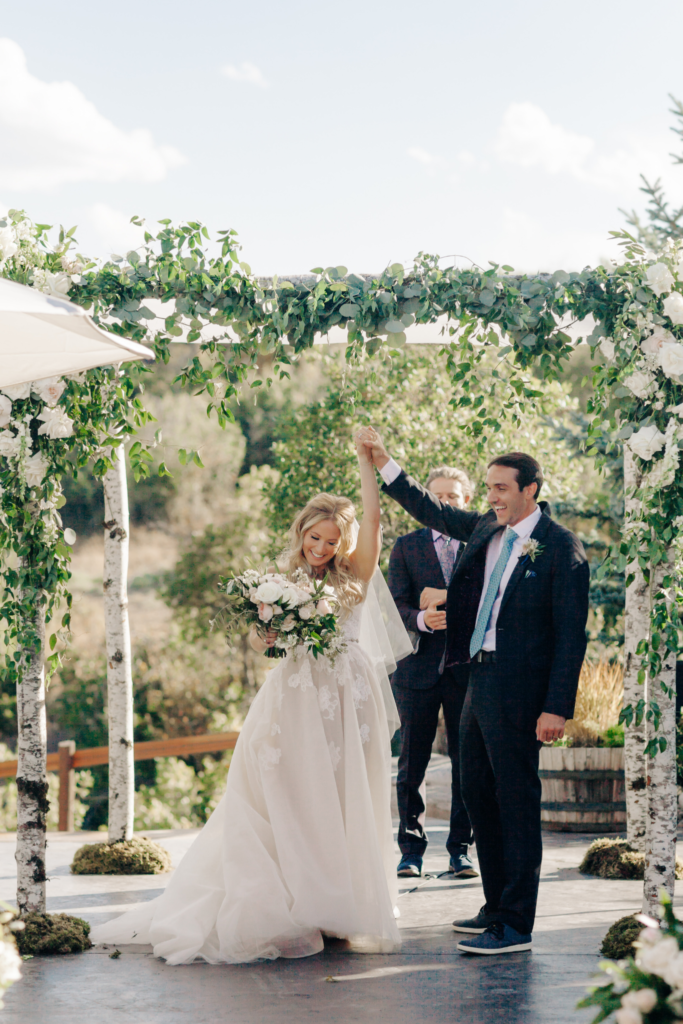 A woman dressed in a white wedding gown. She has blonde curly long hair, has a big smile on her face and carries a bouquet in one hand. Her other hand is holding the grooms hand lifted in the air. Celebrating their non-traditional wedding. The male groom has black hair and wears a black suite and a coral colored tie.