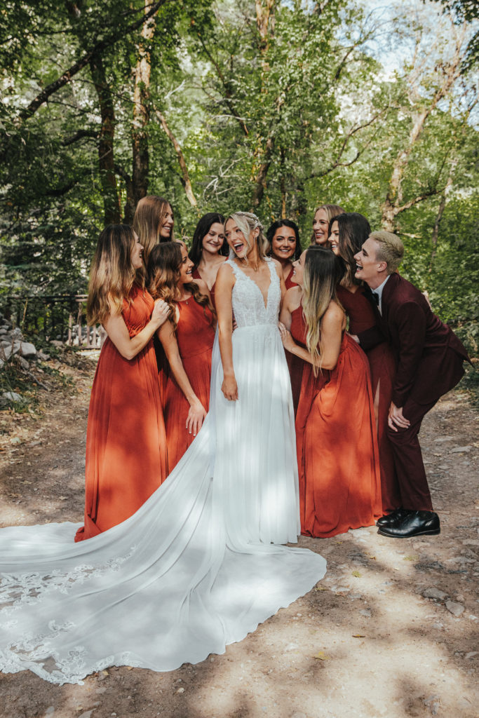 A bridal party of nine people admire the bride. They are crowded around her and the bride's train is splayed out on the ground. Everyone has big smiles.